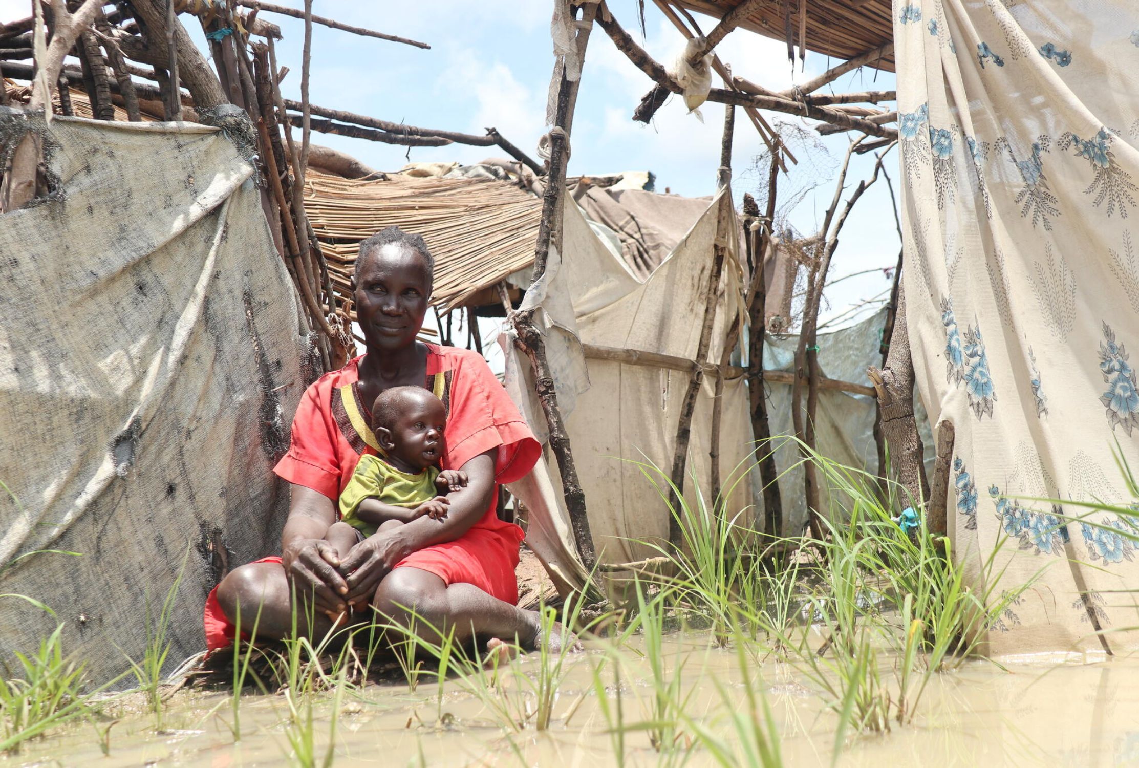 South Sudanese mother and baby sat in front of their flooded makeshift home.