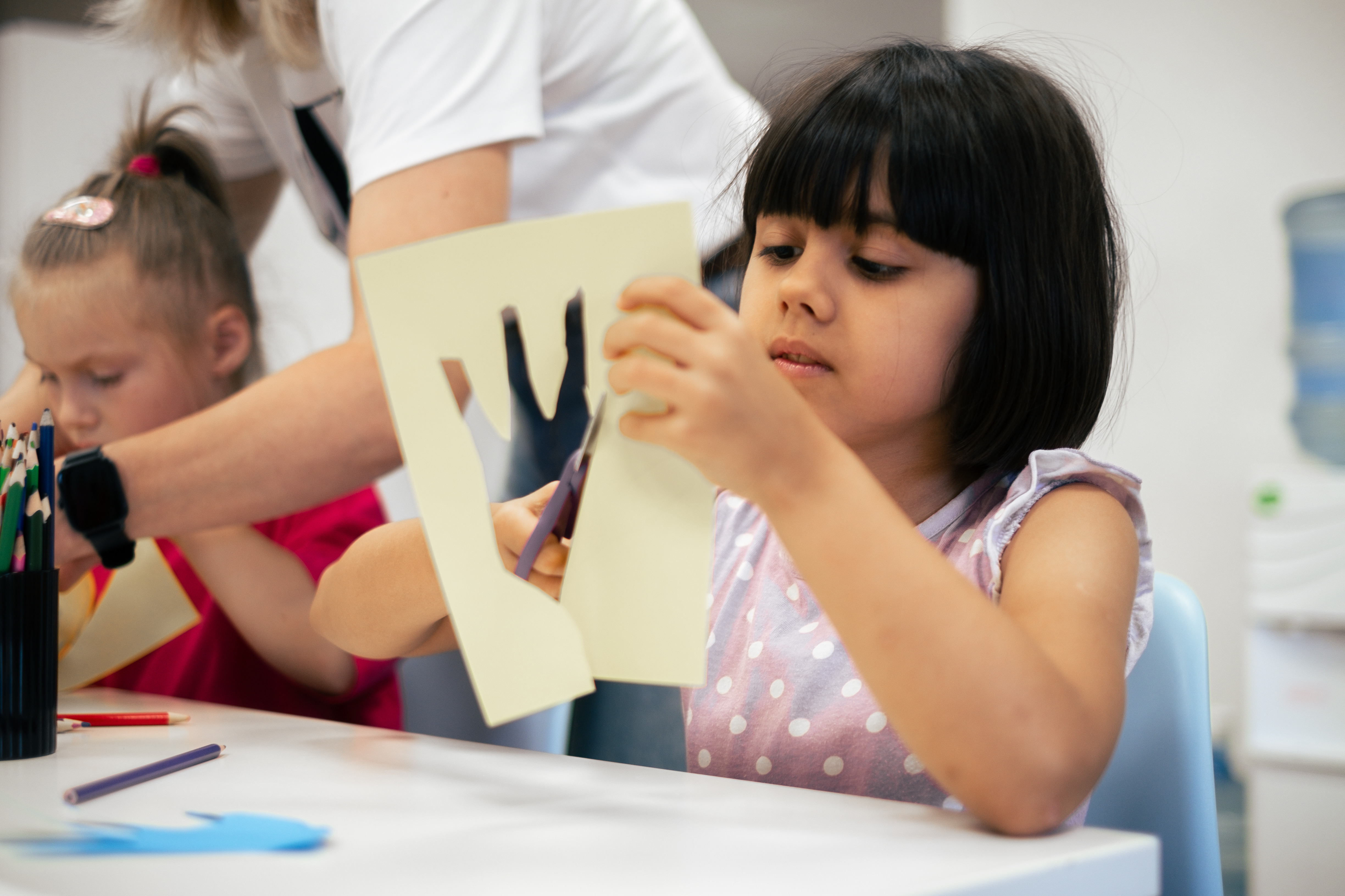 Two girls are cutting paper under guidance from an adult 