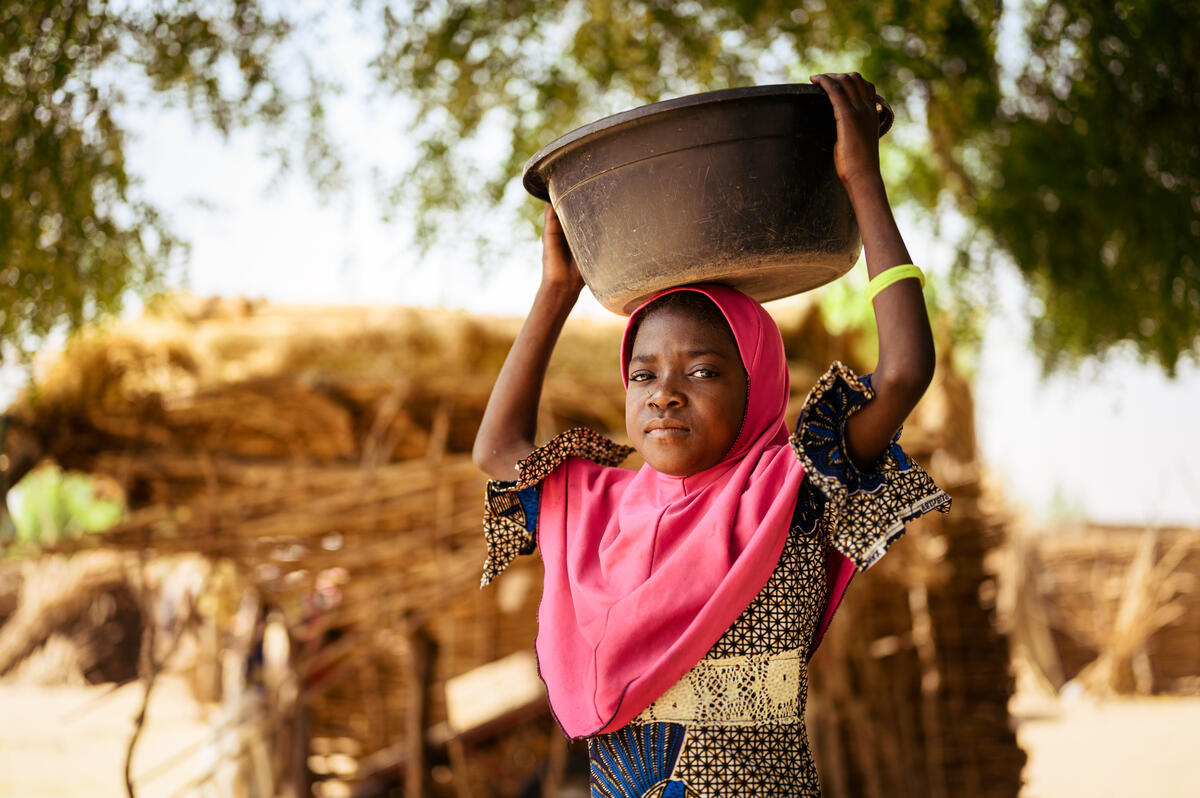 A girl carrying a water container on her head looks directly into the camera
