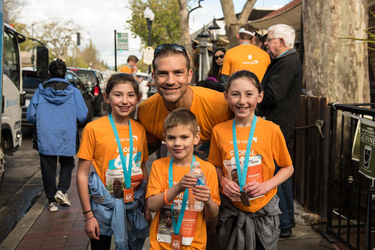 Man in the USA and 3 children smile, wearing orange World Vision tshirts and blue medals after completing the Global 6K race