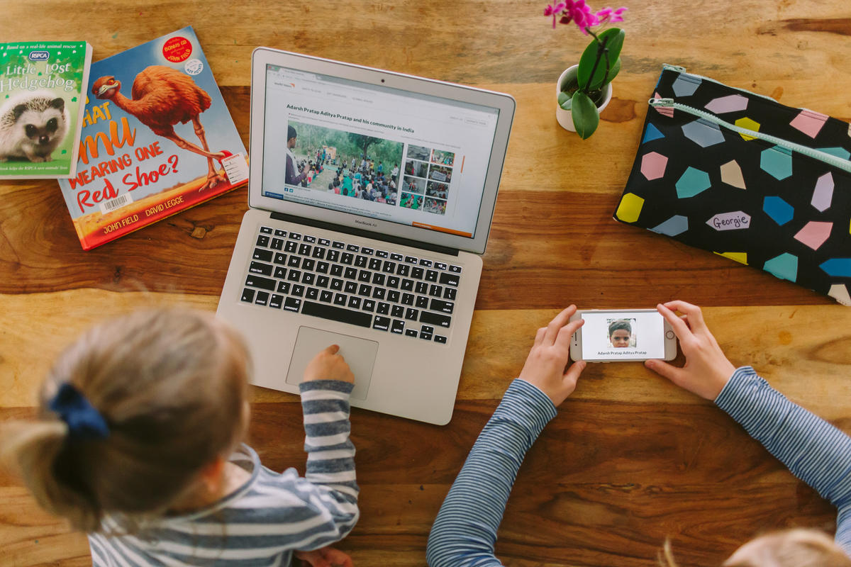 Child sits at a laptop while an adult holds out a mobile