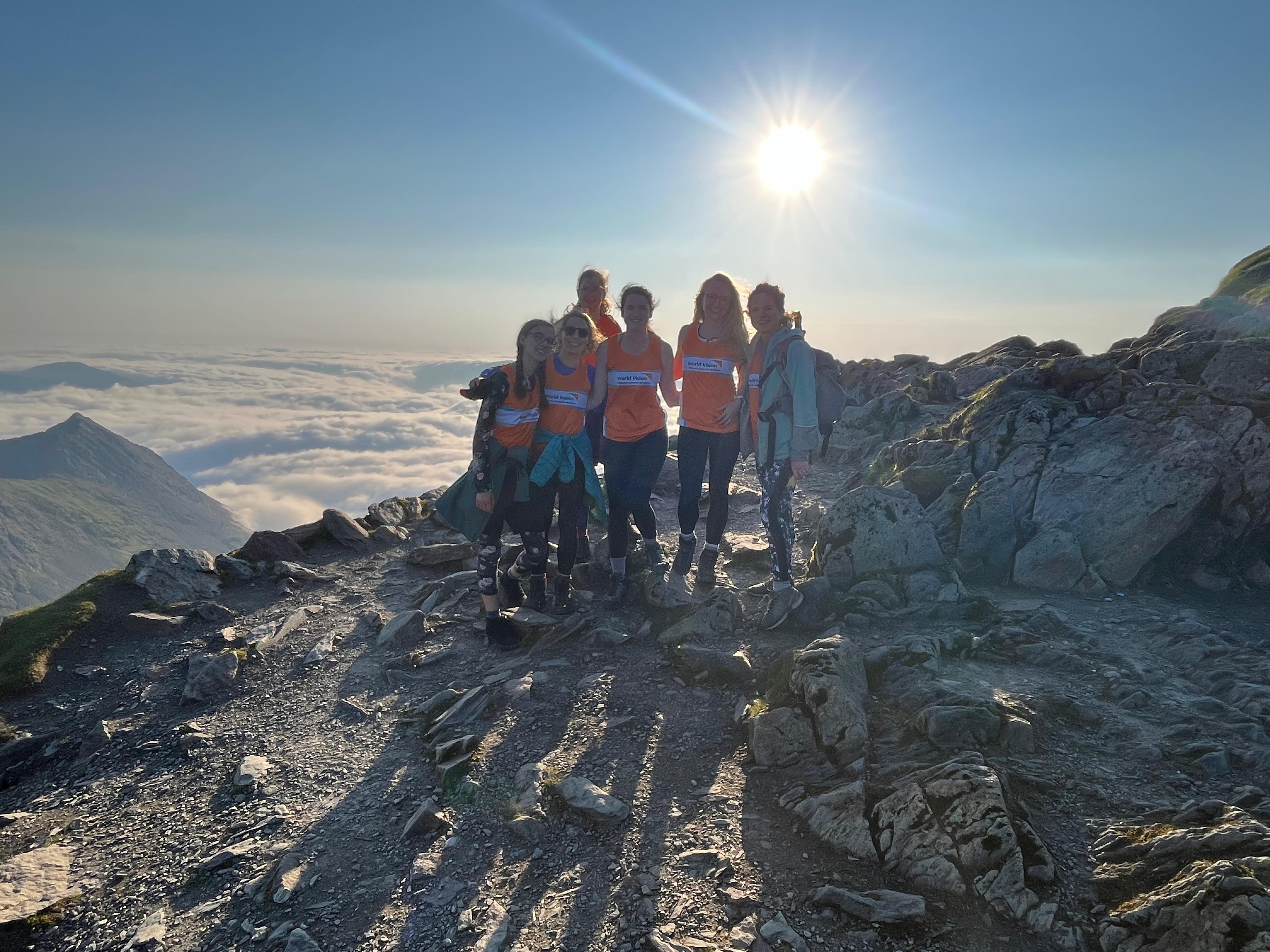 A group of 6 friends pose for a photo on Snowdon, the sun is visible behind them.
