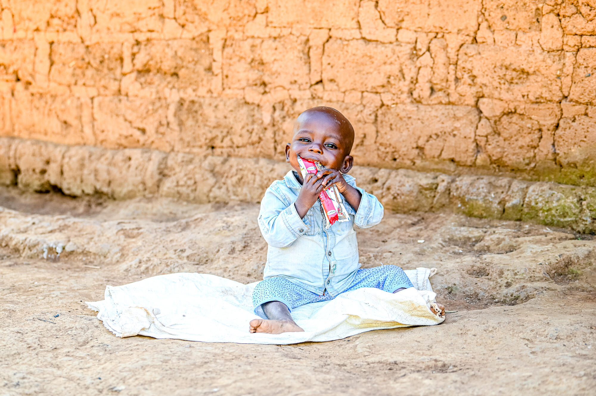A small boy, Geordy, sits outside on a blanket eating from the Plumpy'nut paste packet that has helped him recover from malnutrition in DRC.