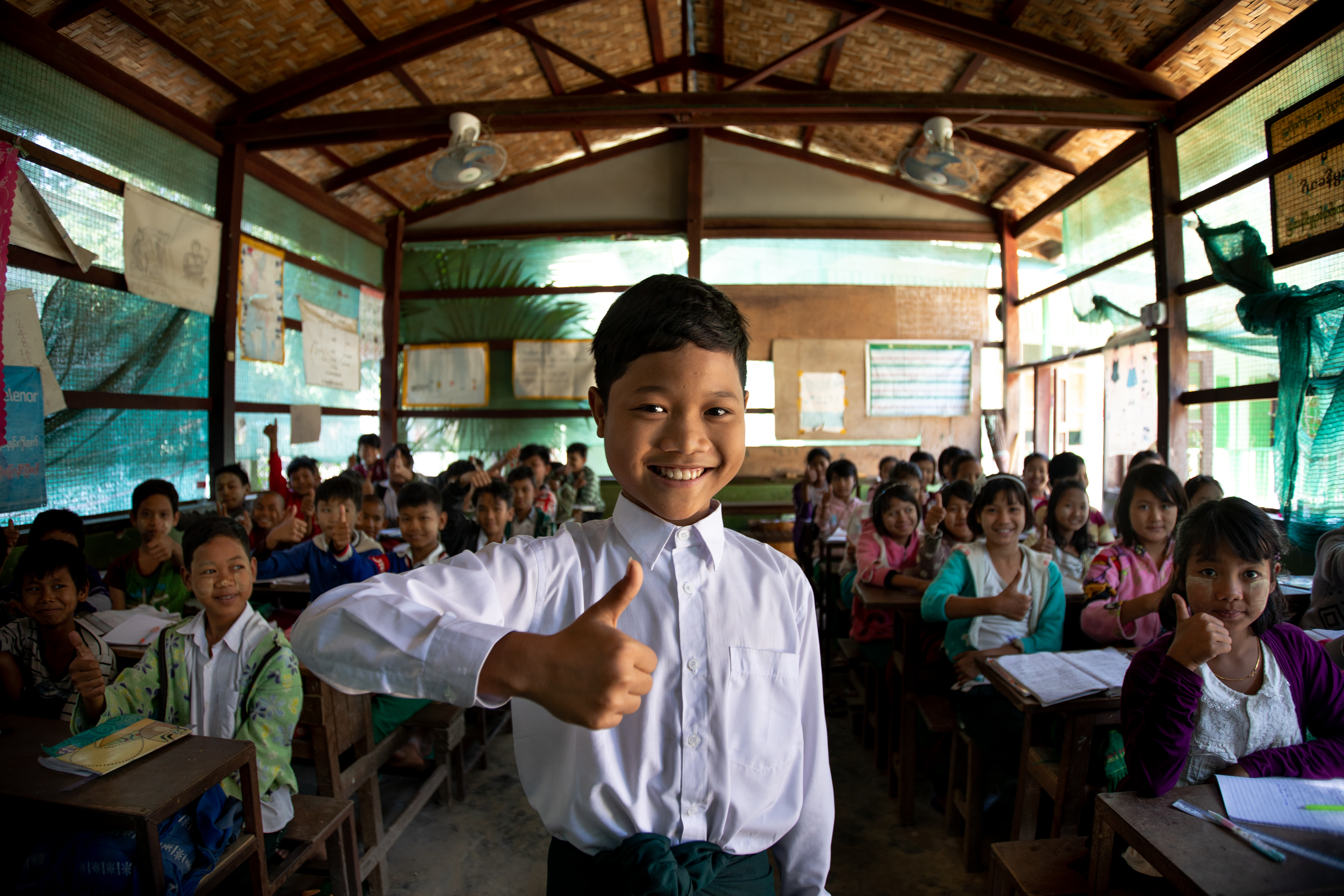 A boy at the front of class giving the thumbs up and smiling with other children sitting behind their desks