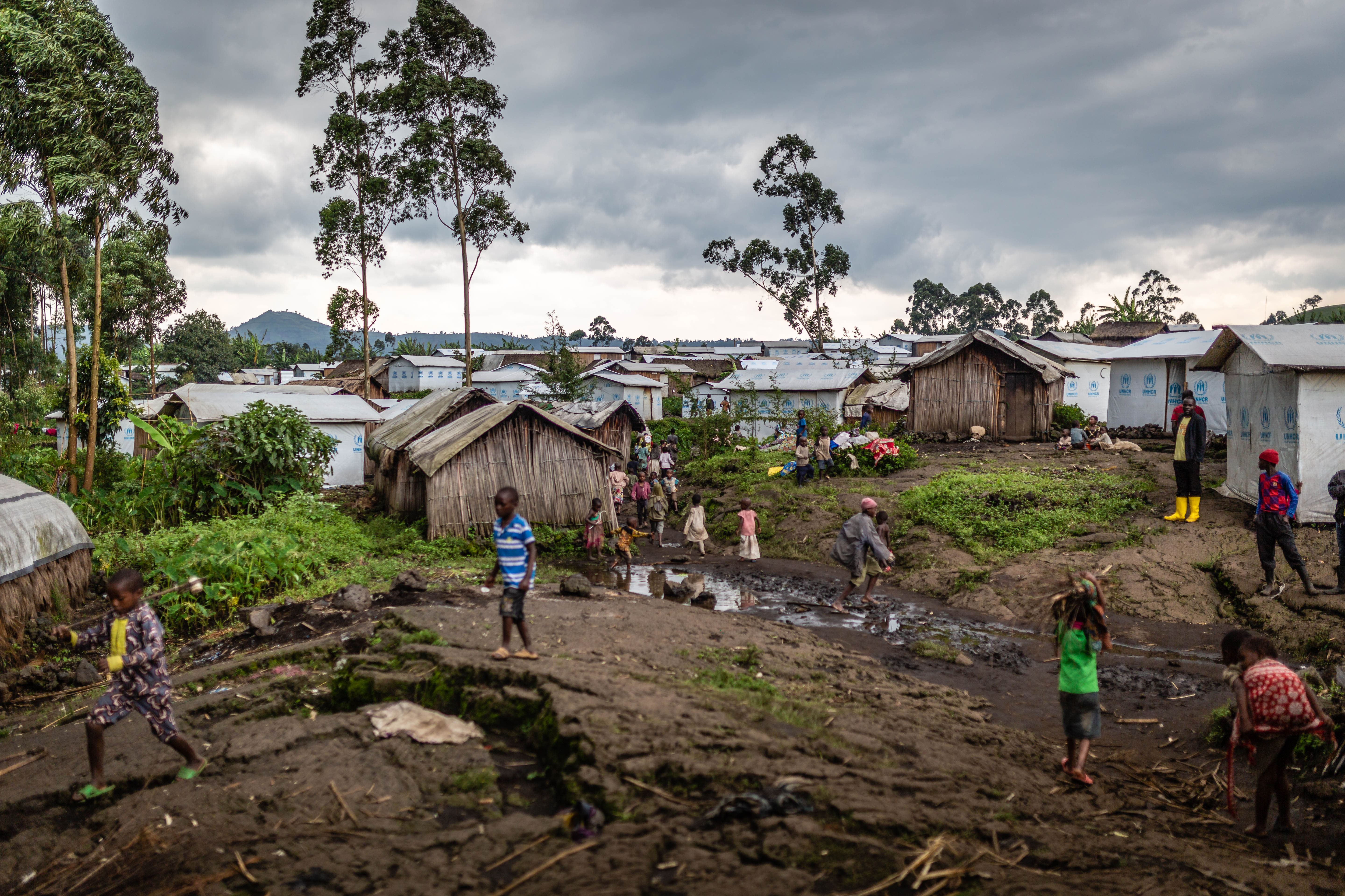 A view of wooden homes and shacks in DRC. The sky is cloudy and children play in the mud outside of the homes.