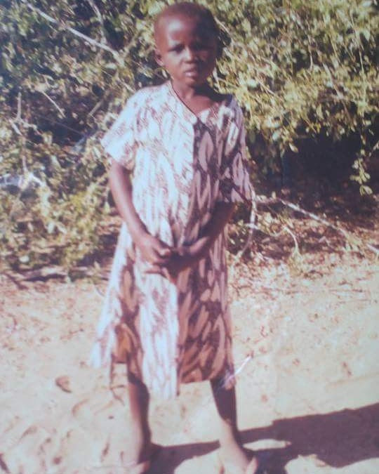 Pauline, as a little girl, poses for her first sponsorship photo in a patterned dress, with scrubland behind