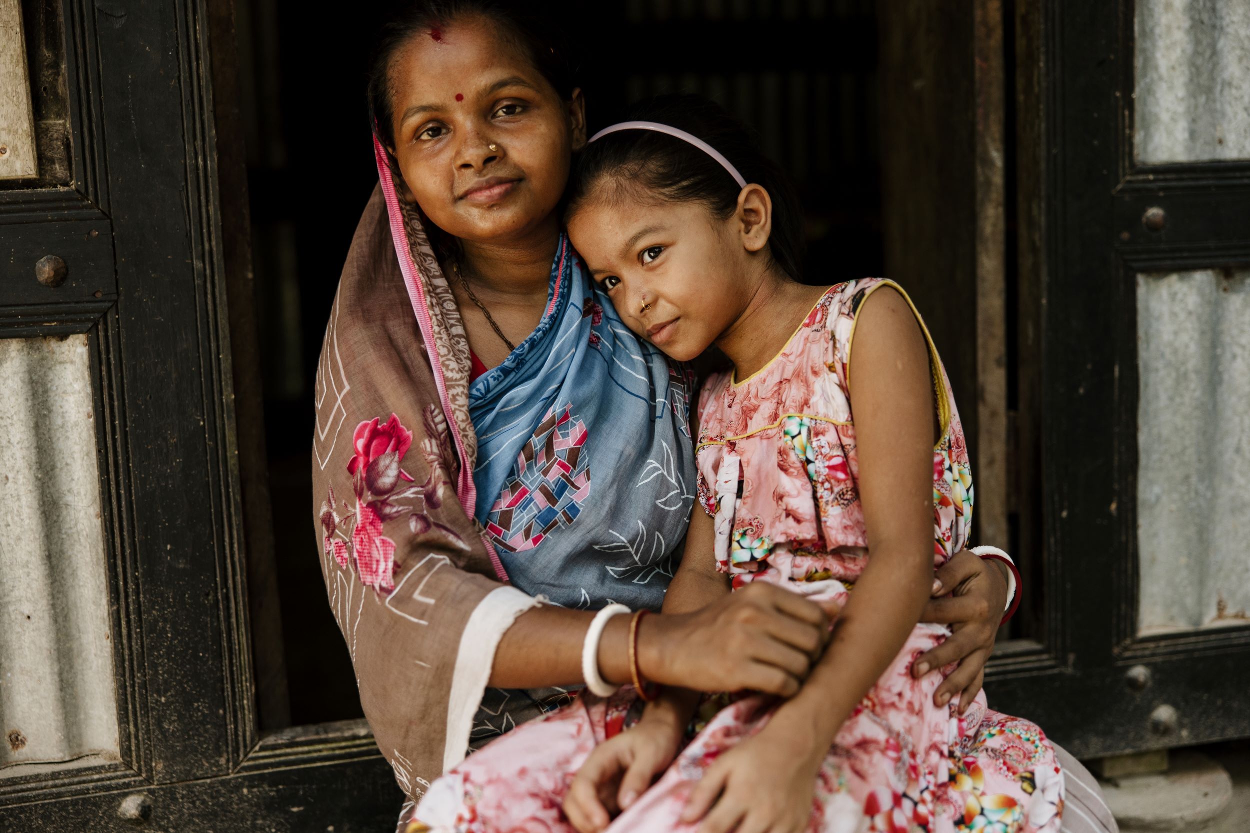 Bangladeshi girl wearing pink dress, sitting on her mother's lap