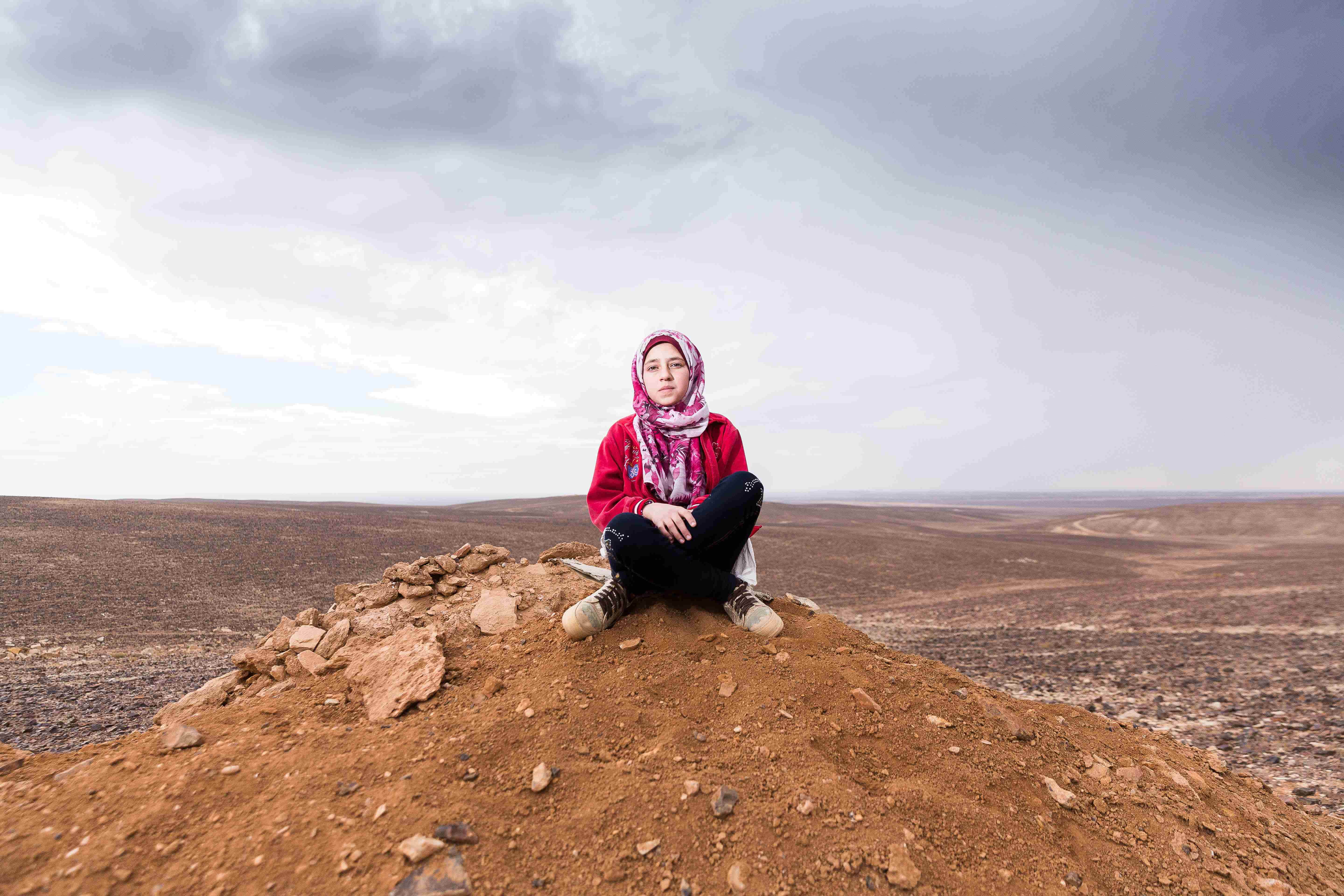 A Syrian girl sits on top of a hill, with blue sky and clouds behind her