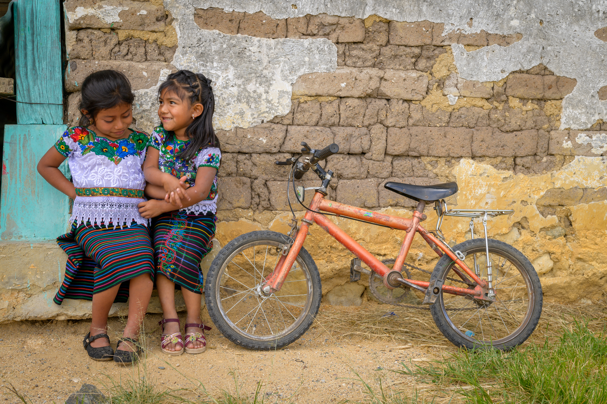 Two young girls sit together outside a house, with bicycle leaning against the wall next to them