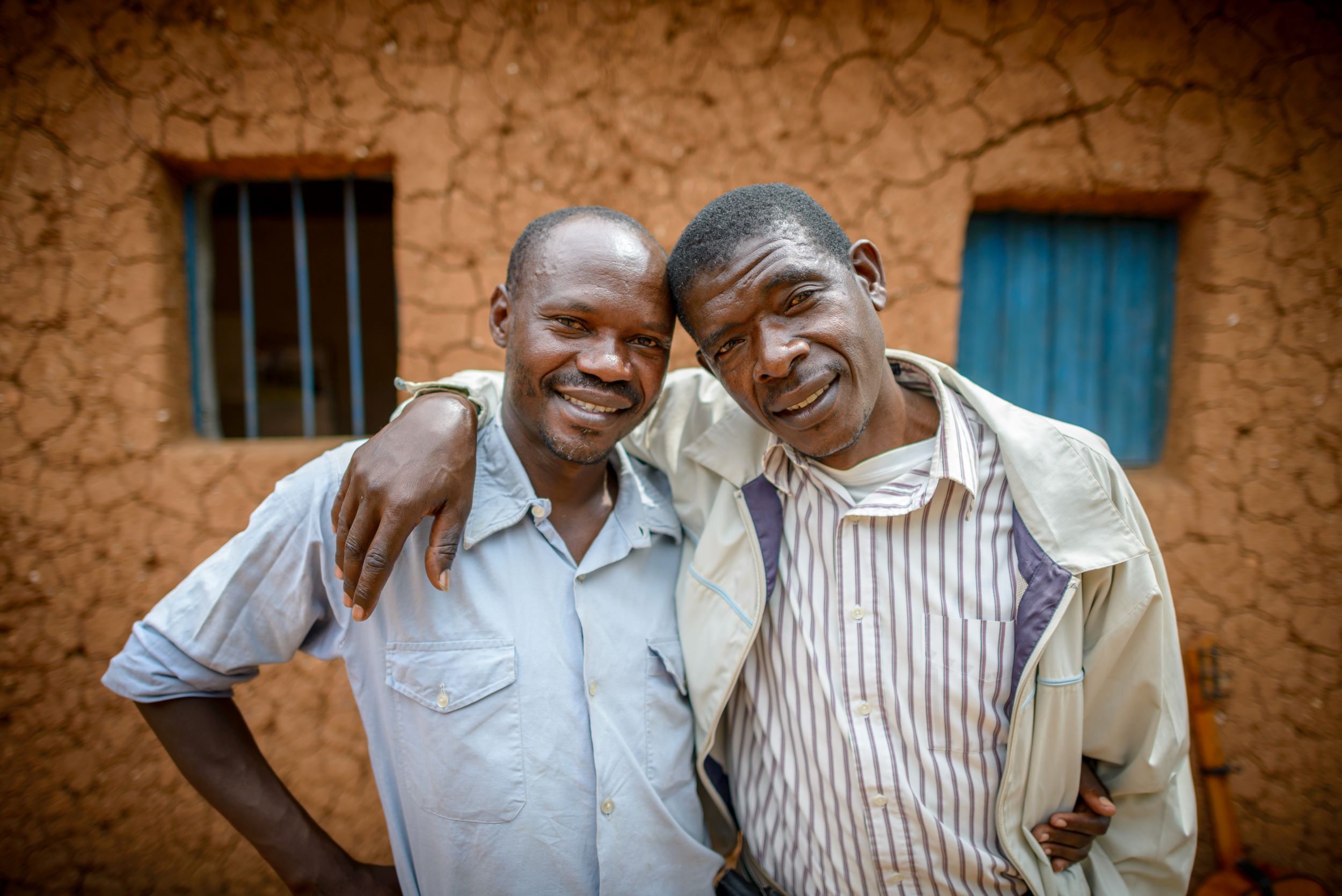 Two smiling Rwandan men with arms around each other in front of a building