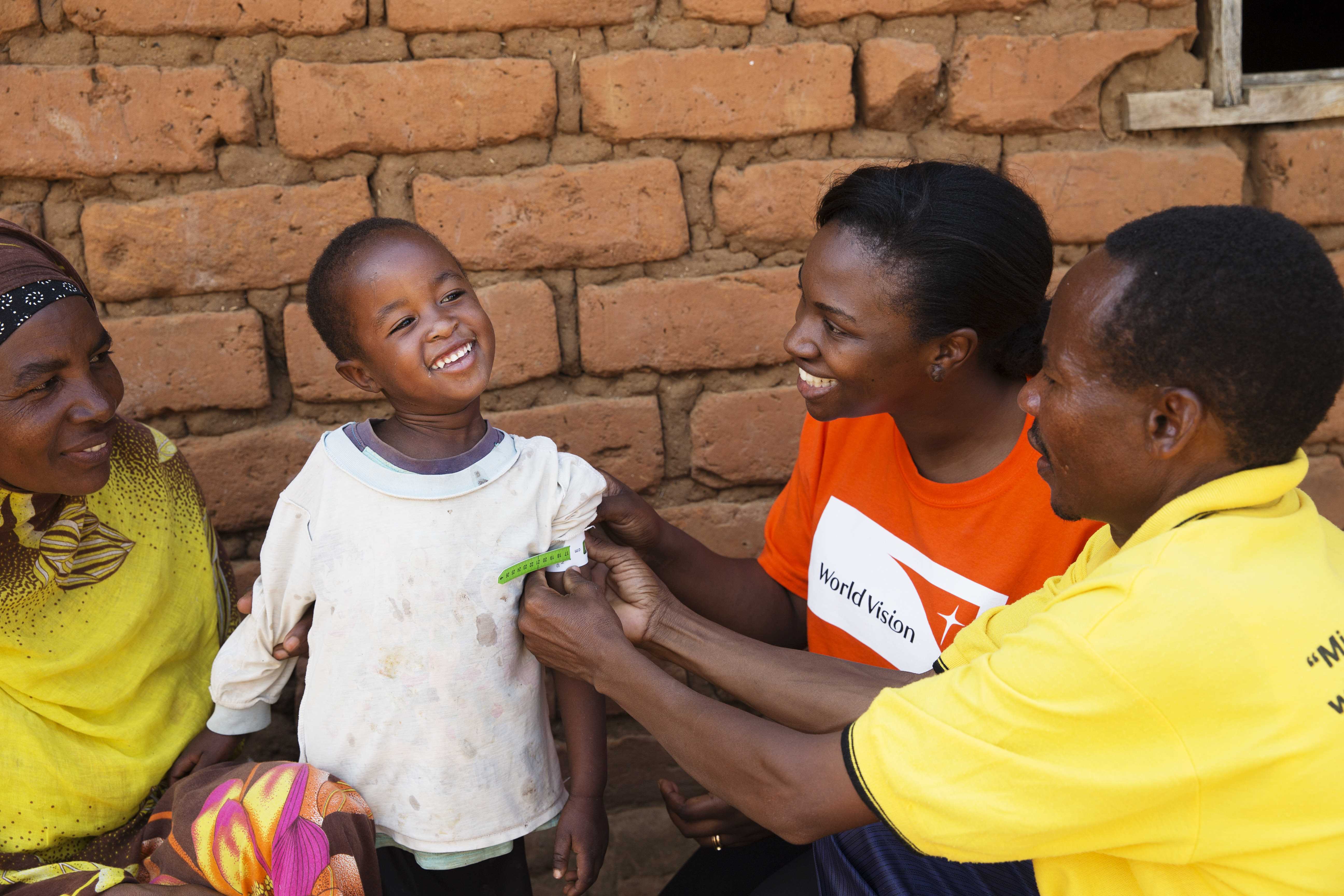 Child in Tanzania gets checked for malnutrition, with three adults dressed in yellow and orange tops around them
