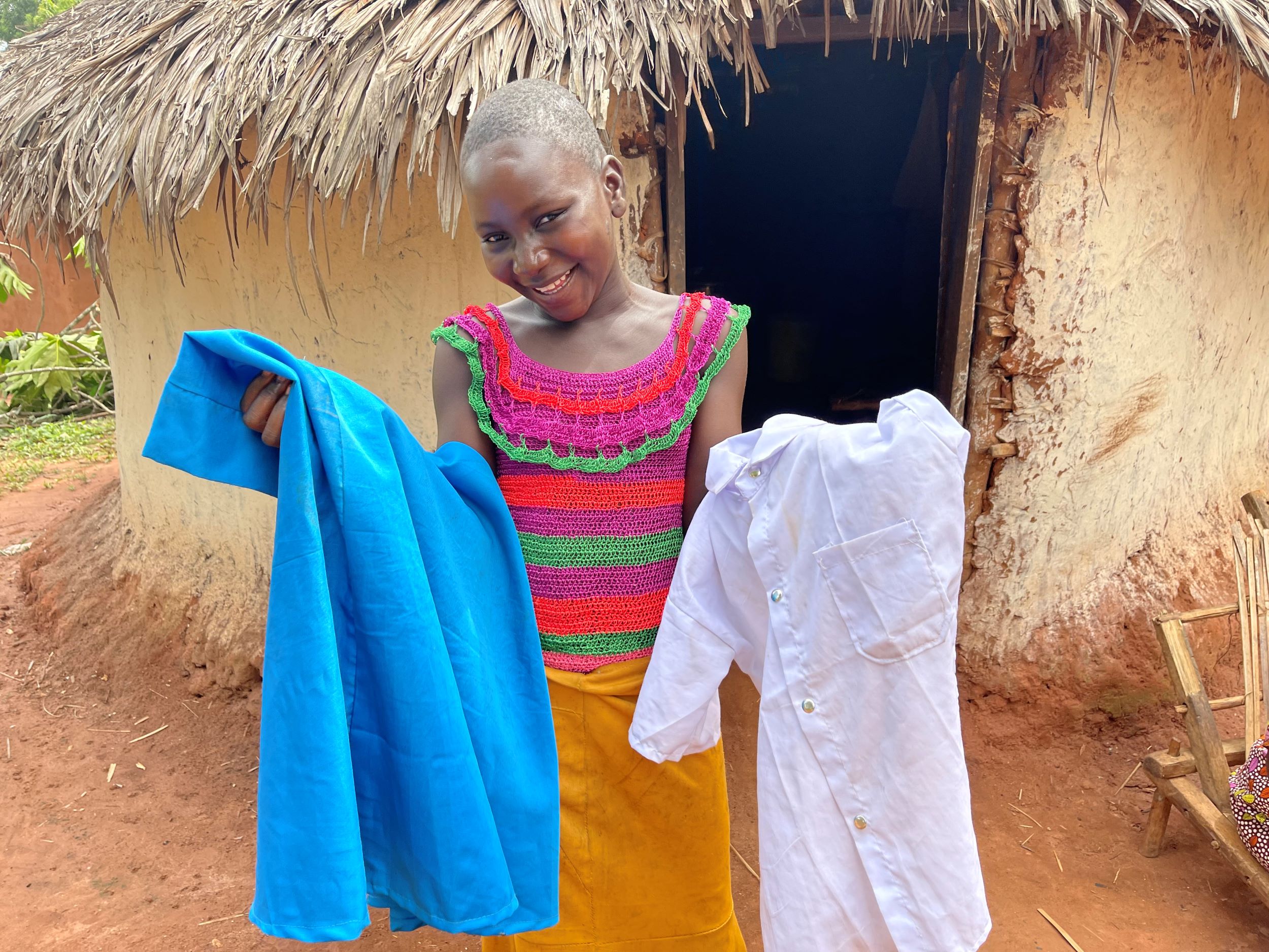 Alt: A young girls stands holding her new uniform – a blue dress and white shirt