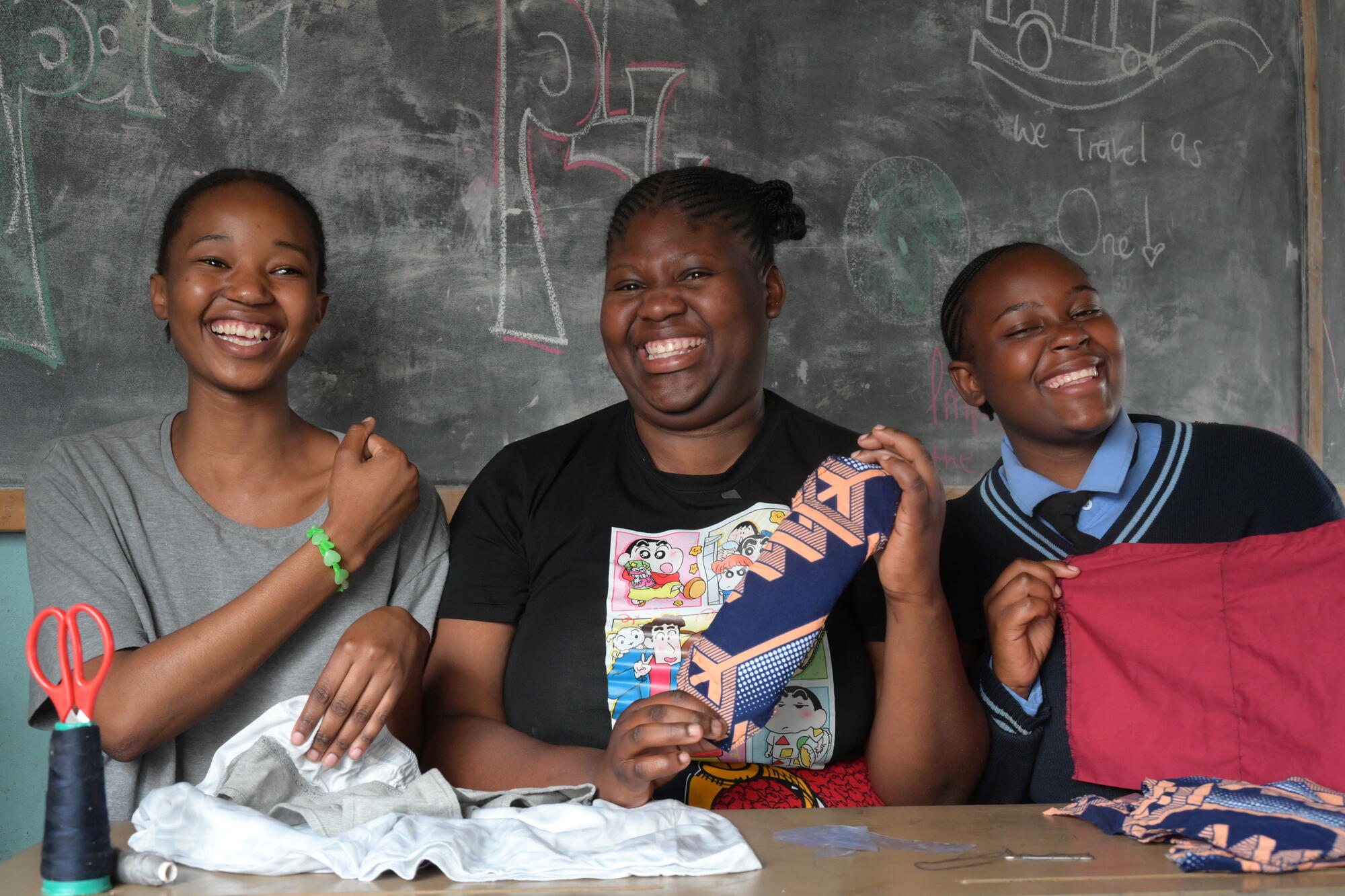 Three women smile to camera in a classroom holding up materials they're using to make reusable sanitary pads
