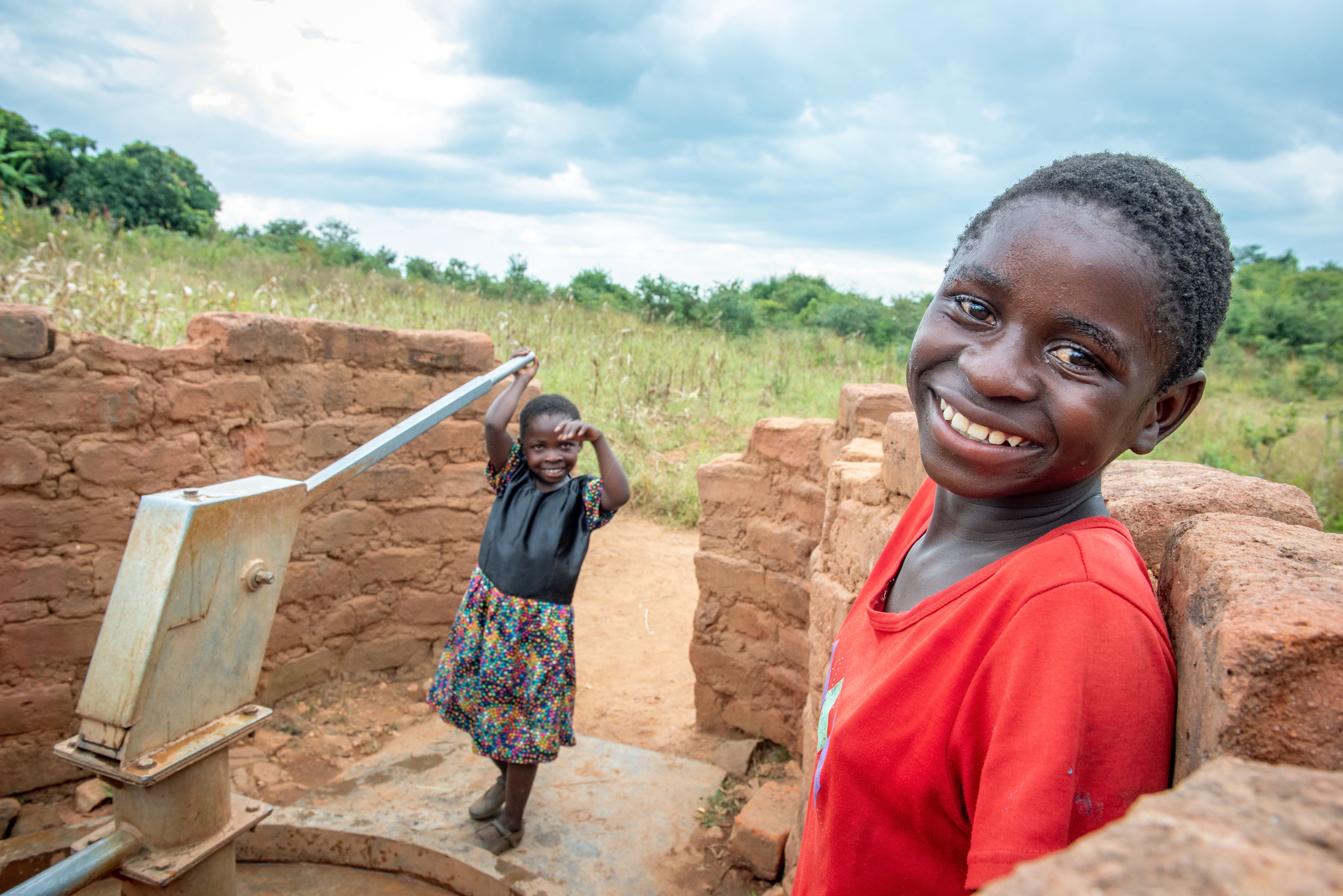 Debby, 7 using a water pump with her friend Brendah, 10 in the foreground looking to the camera