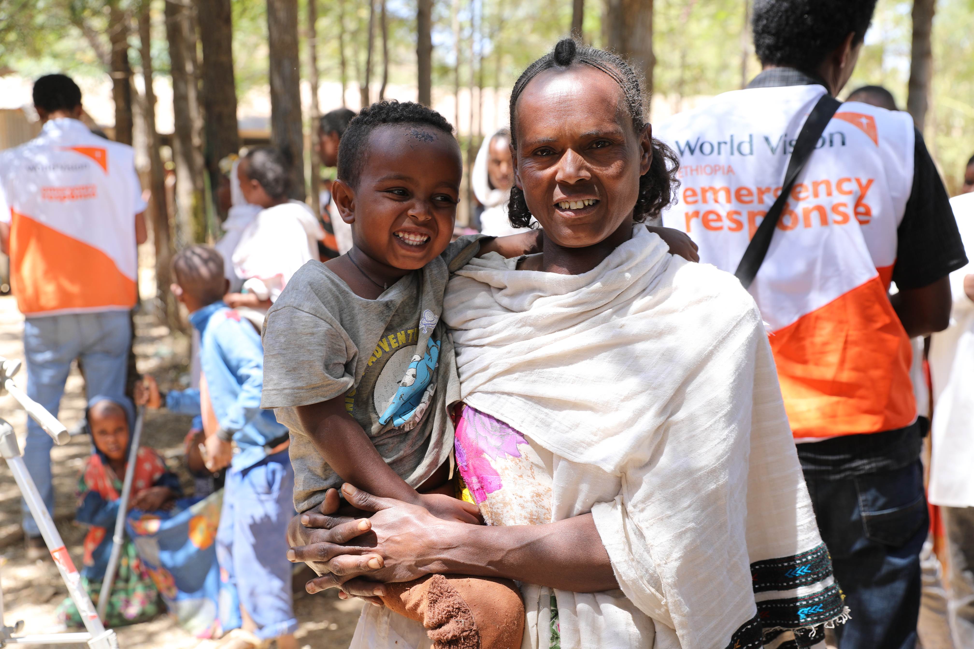 Woman stands in a crowd, holding a child, with emergency response teams standing behind