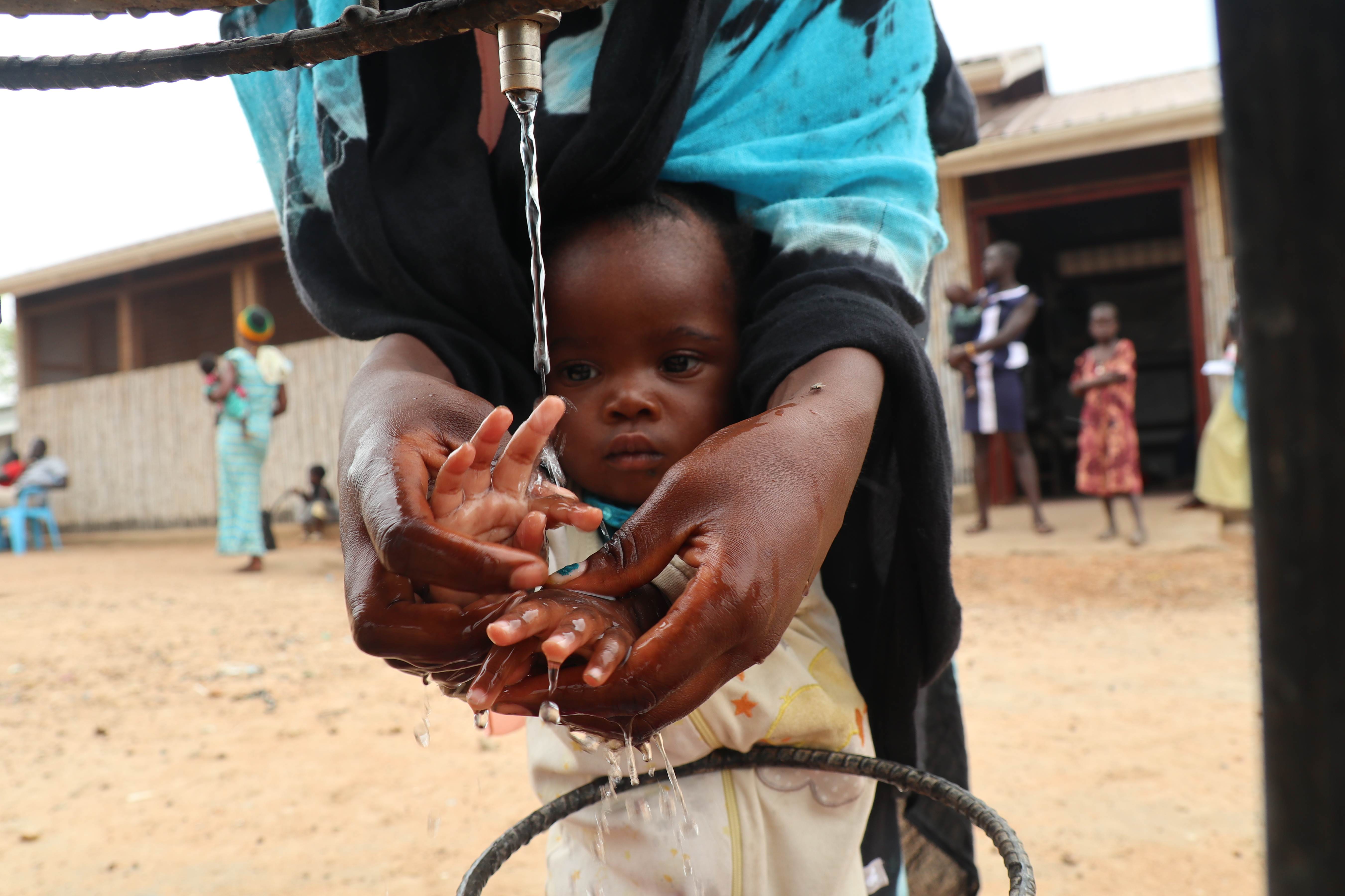 Infant from South Sudan washing their hands with their mother helping from behind them