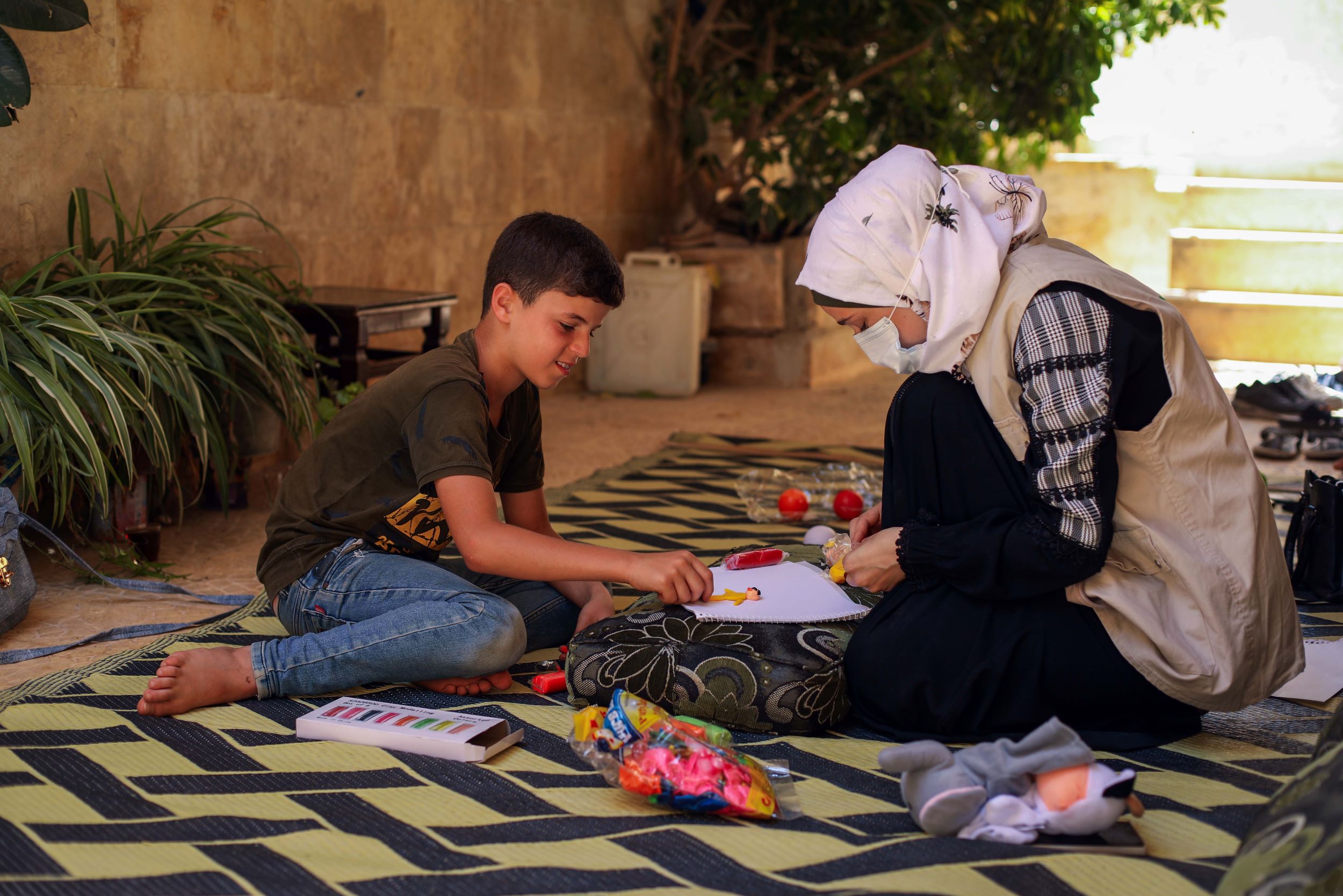 Young Syrian boy sitting on a carpet with a World Vision staff member