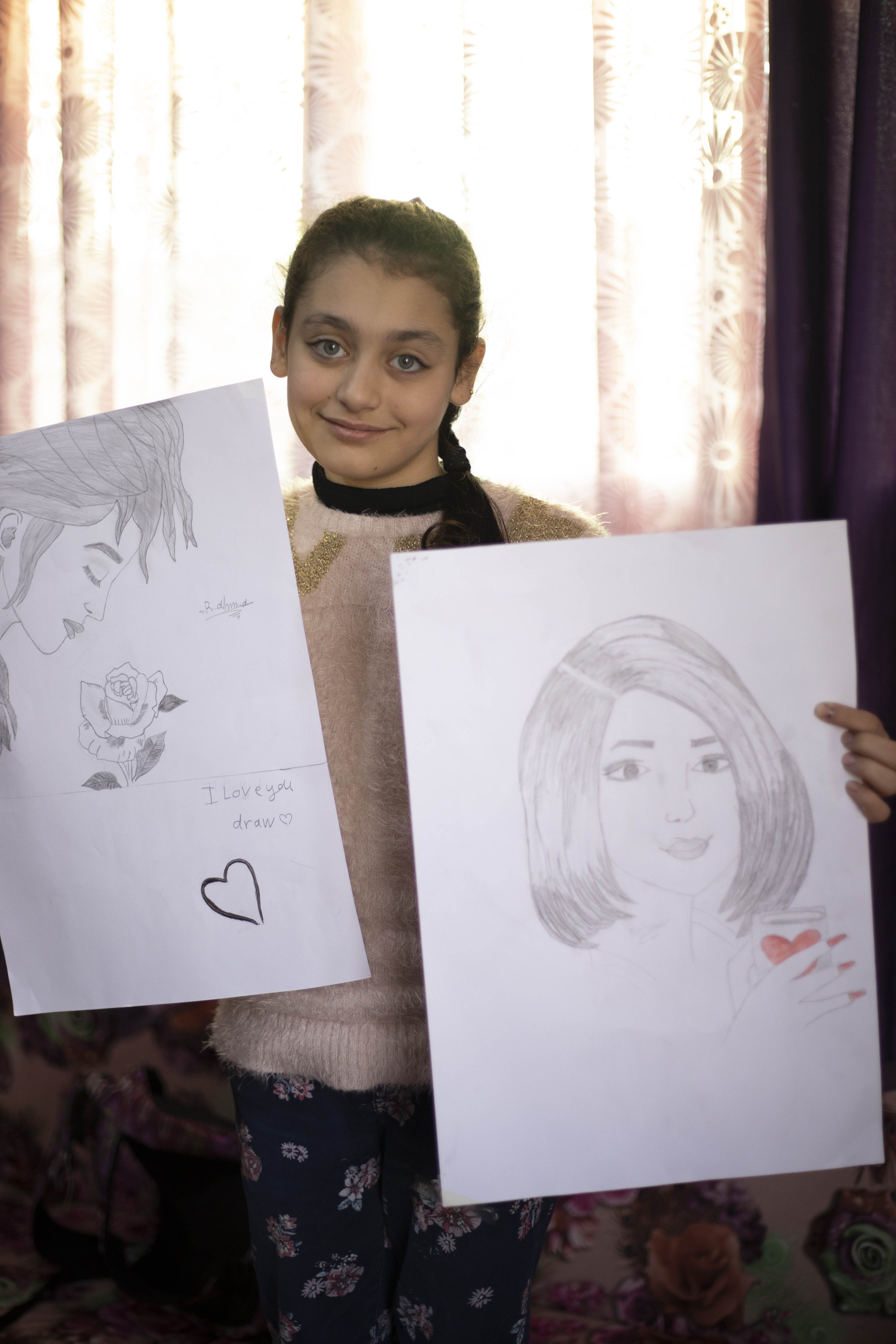Smiling 10 year old Rahma holding up two of the portraits of women's faces that she's drawn